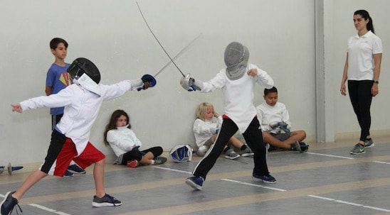 Two kids fencing at summer camp as their fencing camp counselor and other campers look on.
