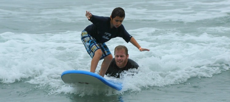 Boy receving surfing instruction from his camp counsleor while enjoying his Los Angeles surf camp experience at summer camp.