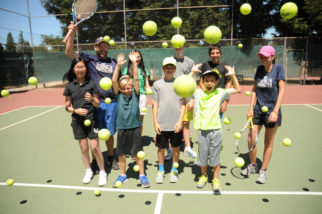 JCC Maccabi Sports Camp kids with counselors standing on tennis court throwing tennis balls into air and laughing.