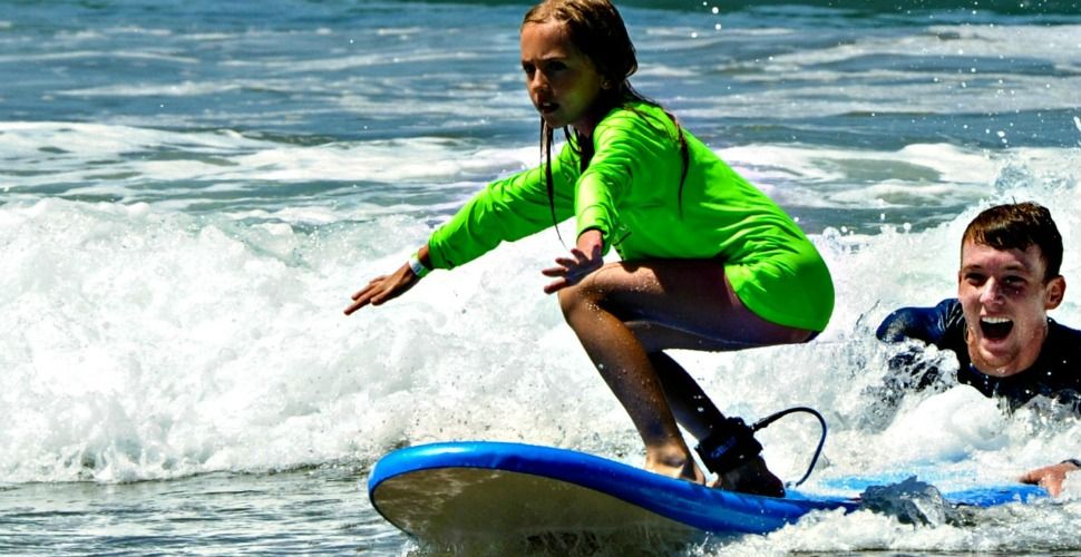 Kahuna camper surfing in the ocean with camp counselor holding her surfboard for support at Aloha Beach Camp, Los Angeles