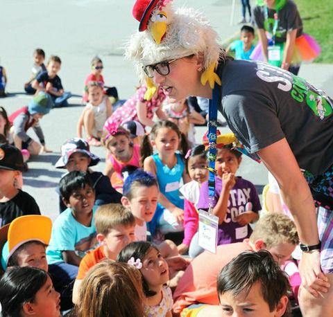 Female camp counselor wearing a funny rubber chicken hat while standing up and bending over to talk to her group of young campers as they listen intently.