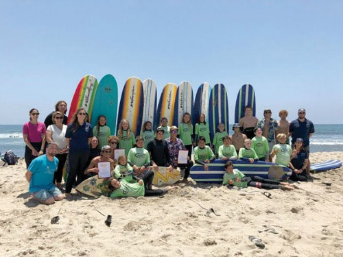 Large group of campers sitting on beach with surfboards behind them in the sand.