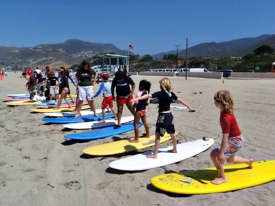 Campers standing on surfboards surfboards on the sand getting a group surfing lesson at Aloha Beach Camp in Los Angeles.
