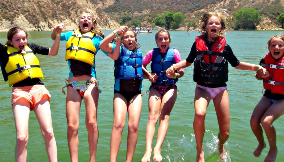 6 girls wearing lifejackes and holding hands, jumping into Castaic Lake at Aloha Beach Camp