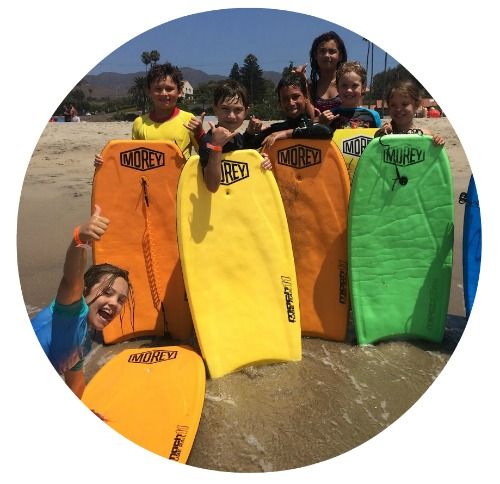 6 kids from Santa Monica standing together with orange, yellow, blue and green boogie boards at the ocean's edge at Aloha Beach Camp