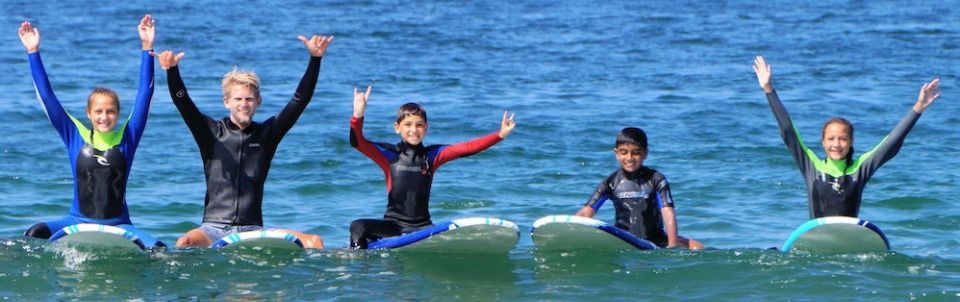 Four kids and their camp counselor sitting on their surfboards in the ocean with their hands up in the air.