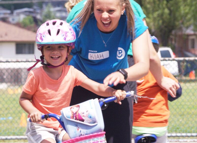 Female camp counselor smiling while teaching a young camper how to ride at bike at Pedelheads summer day camp.