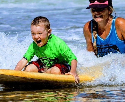 Young boy riding a surfboard on his knees with a big smile on his face while his surf camp instructor holds the back of his surfboard for support.