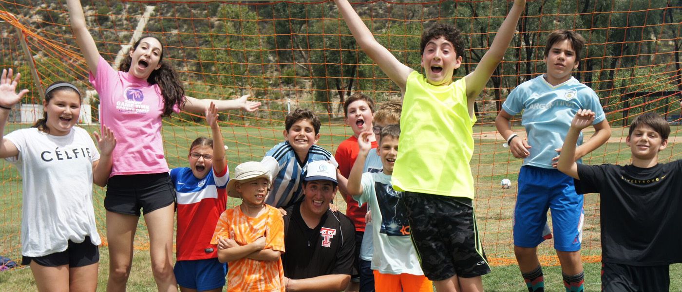 Group of kids with their camp counselor jumping and smiling while standing on a ball field in front of a soccer goal at Saken Sports Camp in Los Angeles.