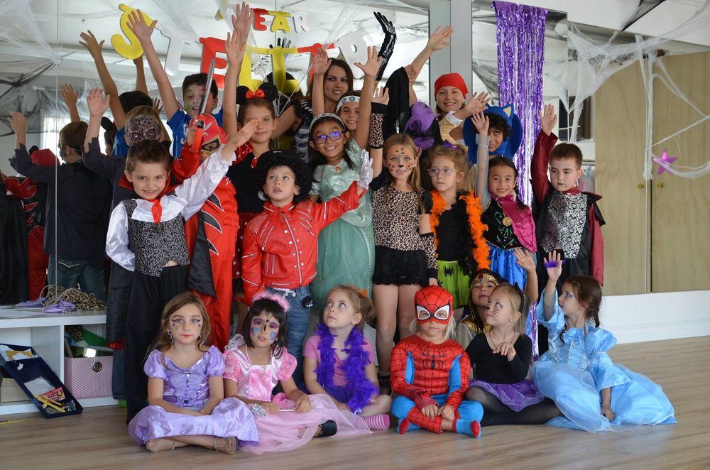 Huge group of young children in colorful Halloween costumes posing for a group picture at Stellar Dance Studio with their two adult dance camp instructors.