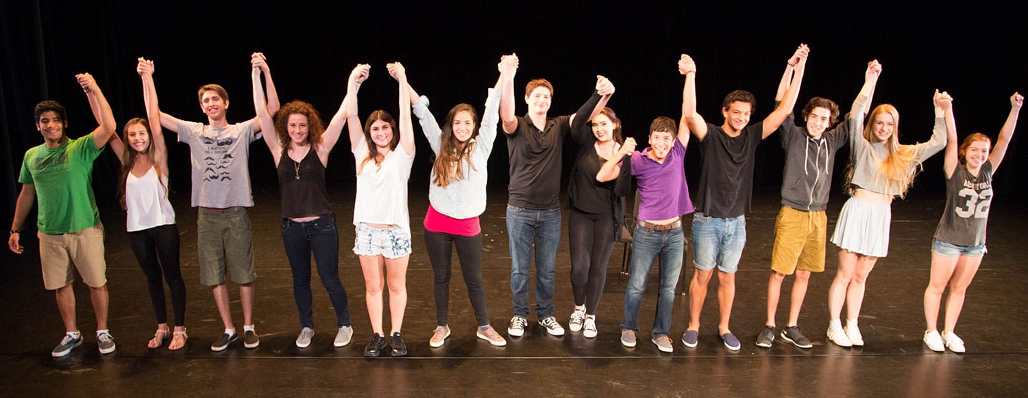 13 high school kids holding hands high over their heads about to take a bow on stage after completing a performance at SOCAPA, School of Creative and Performing Arts Summer Camp