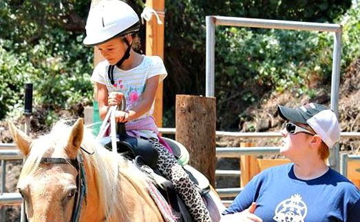 Female camper enjoying the horseback riding activity while receiving supervision and riding instruction from her Camp Counselor at Tumbleweed Day Camp.