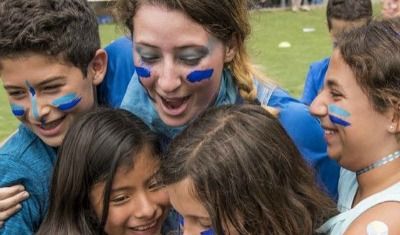 4 kids and their camp counselor with their faces painted blue playing games together on an outdoor grass ballfield at summer camp.